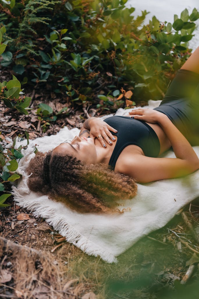 A woman with curly hair meditates on a white towel amidst greenery outdoors, embracing nature.