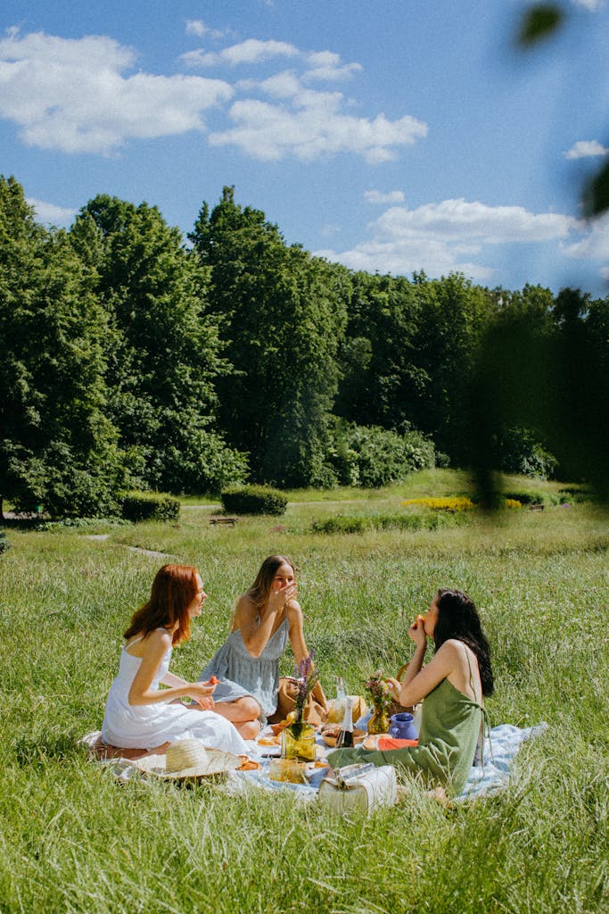 Three women enjoy a serene picnic in a sunny park with lush greenery.
